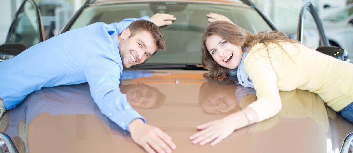 Happy couple embracing car in dealership and looking at camera.